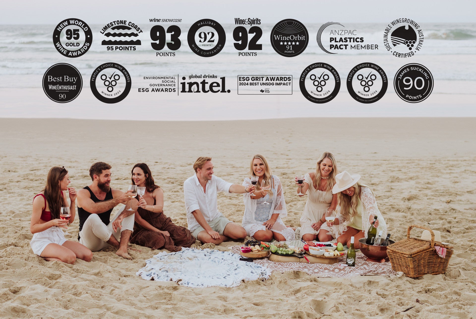 Beach picnic scene with a group of friends sharing wine and snacks on the sand, with ocean waves in the background. Award and certification logos are displayed across the top, including wine ratings, sustainability certifications, and ESG awards.